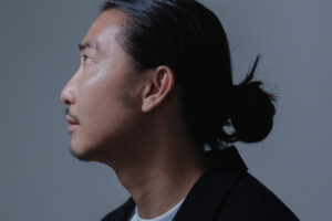 Dramatic Headshot Side profile of a man with a topknot hairstyle, wearing a black jacket.