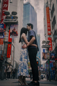 Authentic Couple Photography A couple embracing in a busy Shibuya street with tall buildings and colorful signs in the background.