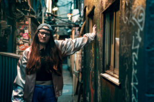 Cinematic Outdoor Portrait A woman in a shiny jacket and glasses stands in a narrow Shinjuku alleyway, touching a wall with graffiti.