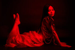 Portrait with RGB Light A Japanese woman in a flowing red dress adorned with beaded necklaces, sitting gracefully with her hands clasped, illuminated by a red light against a dark background.