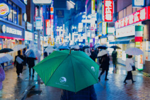 Product Photography for the American Brand Miry Brook A person holding a green umbrella walks through a bustling, neon-lit street in Shibuya at night.