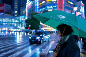 Tokyo Night Scene at Shibuya Crossing by JG Brasseur for the American Brand Miry Brook Person holding a green umbrella at a bustling Shibuya street crossing at night, with city lights and cars in the background.
