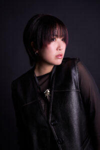 Hong Kong Singer & Artist Kendy Suen A woman with short hair and subtle pink highlights, wearing a sleek leather vest over a dark outfit.