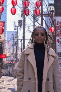 Shibuya Street Fashion by JG Brasseur | Tokyo Lifestyle Street fashion photography by JG Brasseur in Shibuya, Tokyo. A person in a beige sheepskin coat and round sunglasses stands against a backdrop of red lanterns and urban scenery. Captures the vibrant energy and style of Tokyo's streets.
