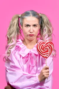 Pink Pop & Heartbreak: Conceptual Fashion Portrait with French Singer AUDE A high-fashion portrait of a woman with silver-blonde pigtails and a moody expression, wearing a ruffled pink blouse and holding a large red-and-white heart-shaped lollipop against a vibrant pink background.