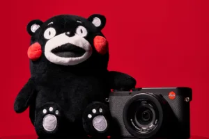 Kumamon & Leica: Red Dot Chili Peppers - Studio Product Test Shoot A black Kumamon plush mascot sitting next to a black Leica digital camera against a vibrant solid red background.