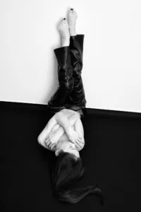 Minimalist Monochrome Study Black and white studio portrait of a woman lying on a dark floor with legs raised against a white wall, wearing black leather trousers and crossing her arms over her chest.